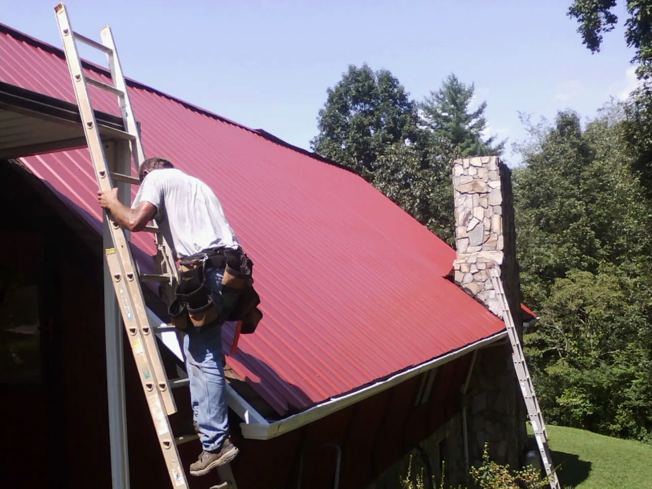 Roofer on a ladder beside a red metal roof and stone chimney — Soffit & Fascia Repair by Roofing Brothers in Nelson